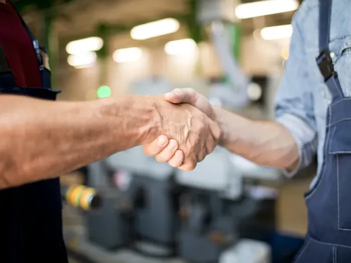 Close-up of two workers shaking hands in a factory or workshop setting, symbolizing teamwork and trust